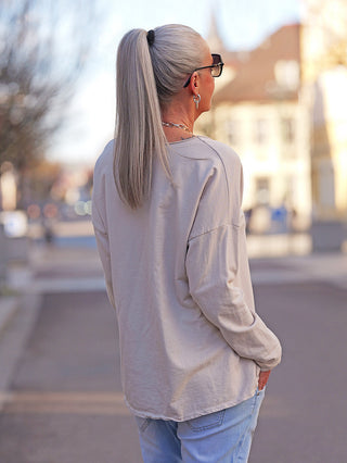 Eine Person trägt das OFF#DLY LS Mia beige Longsleeve mit V-Ausschnitt und lockerem Schnitt, kombiniert mit Jeans und Accessoires, im Sonnenlicht auf einer ruhigen Straße.