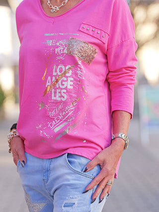 Nahaufnahme des Oberkörpers, der das OFF#DLY LS Mia pink Longsleeve trägt, mit gold-silbernem Schriftzug Los Angeles, Ziernieten auf der Brusttasche und silbernen Accessoires, kombiniert mit hellen Jeans.