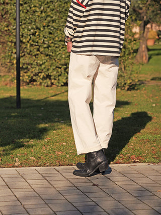 Person trägt OFF#DLY Joggpant Melania creme, locker geschnittene weiße Hose, knöchellang, aus Baumwollstretch, mit seitlichen Taschen und umgeschlagenen Säumen, kombiniert mit gestreiftem Shirt und schwarzen Lederstiefeletten.