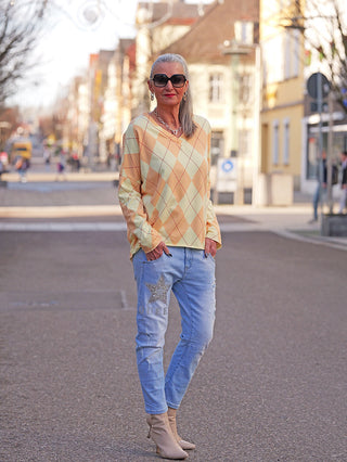 Frau trägt OFF#DLY LS Karo yellow/orange Pullover mit argyle-Muster, entspannter Schnitt. Kombiniert mit Jeans und Stiefeletten, betont der Pullover lässige Eleganz für vielseitige Alltagslooks.
