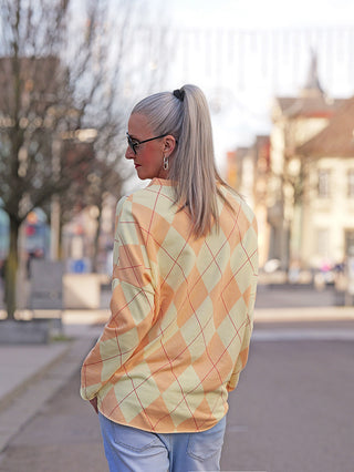 Person von hinten mit silbergrauem Pferdeschwanz trägt OFF#DLY LS Karo yellow/orange: locker fallendes Longsleeve mit argyle Muster, V-Ausschnitt, kombiniert mit hellblauen Jeans auf einer Stadtstraße.