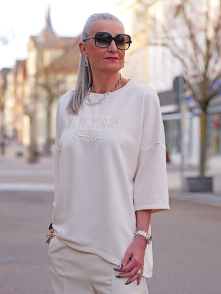 Person trägt den OFF#DLY Sweater Shine vanille mit aufgesticktem NEW YORK 5th AVE, kombiniert mit passenden Hosen, großen Sonnenbrillen und silbernen Accessoires, in urbaner Straßenszene.