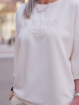 Nahaufnahme des Oberkörpers mit dem OFF#DLY Sweater Shine vanille, der subtil NEW YORK 5th AVE. bestickt trägt. Silberne Kette und Armbänder ergänzen das stilvolle Design des Pullovers.