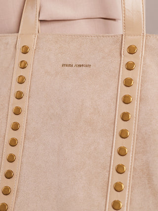 Nahaufnahme einer Regina Schrecker Shopper taupe Tasche in Veloursleder-Optik, mit goldfarbenen Nieten und Ledergriffen mit Nieten. Das Markenlogo REGINA SCHRECKER ist in Goldfolie auf der Vorderseite geprägt.