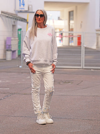 Ältere Frau trägt OFF#DLY Sweat SOHO lightgrey, ein lässiges, graues Sweatshirt mit pinkem Brustmotiv, kombiniert zu Jeans mit Patchwork und weißen Sneakers für urbanen Style.