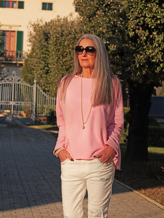 Frau trägt den OFF#DLY Sweat Lotte rose, ein locker geschnittener, rosafarbener Boxy-Sweater mit langen Ärmeln, kombiniert mit weißen Jeans und Halskette, ideal für einen entspannten, stilvollen Look.