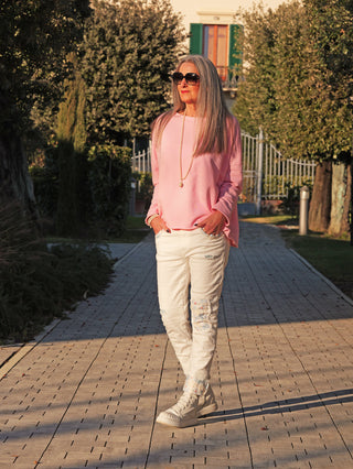 Erwachsene Frau trägt OFF#DLY Sweat Lotte rose, einen locker fallenden, langärmligen Sweater mit Rundhalsausschnitt, kombiniert mit weißen Jeans und Sneakers, auf einem Gartenweg.