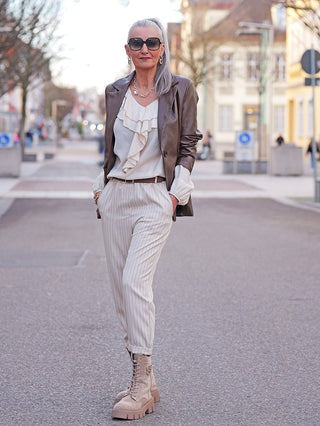 Frau trägt den OFF#DLY Fakeleather Blazer Vasto braun, kombiniert mit einer rüschigen Bluse und beigen Hosen, während sie eine urbane Straße entlanggeht.