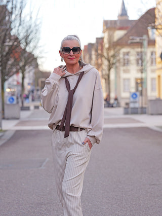 Frau trägt den OFF#DLY Hoody Pescara beige mit auffälligem Krawatten-Detail, kombiniert mit hoch taillierten, gestreiften Hosen, schwarzem Gürtel und oversized Sonnenbrille, stehend auf einer Fußgängerstraße.