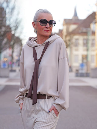 Frau trägt den OFF#DLY Hoody Pescara beige, locker geschnitten mit Kordel im Krawatten-Look, kombiniert mit hoch taillierten, gestreiften Hosen, urbanem Hintergrund leicht unscharf.