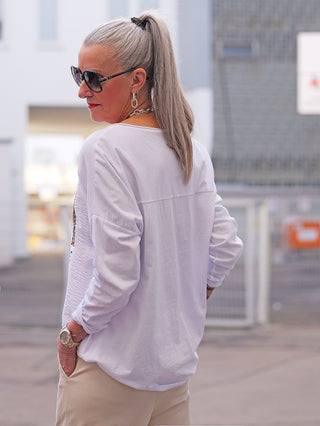 Frau trägt OFF#DLY LS Ella white Longsleeve mit lockerem Schnitt und hohem Tragekomfort, kombiniert mit einer goldenen Uhr und Halskette, stehend in urbaner Umgebung.