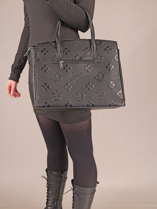 Person trägt ein Sweet Years Shopper 6929 black, eine große, strukturierte Handtasche mit vierblättrigem Kleeblattmotiv, Blumen und kleinen Logos. Die Tasche hat zwei Henkel und eine vordere Reißverschlusstasche.