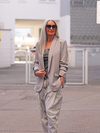 Ältere Frau in OFF#DLY Blazer Diana grey, trägt ihn offen über einem dezent gemusterten Top, kombiniert mit lässigen, weit geschnittenen Hosen; ein elegantes und vielseitiges Statement-Piece.