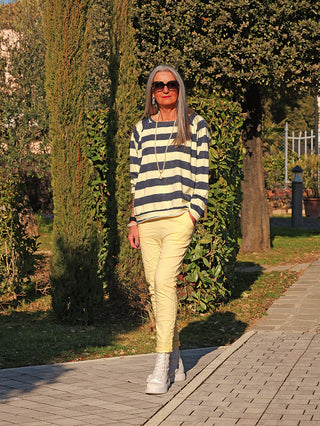 Eine schlanke Frau trägt den OFF#DLY Sweat Lola blue/yellow mit breiten Streifen, während sie im Garten spaziert. Kombiniert mit hellen Hosen und weißen Sneakers für einen lässigen Look.