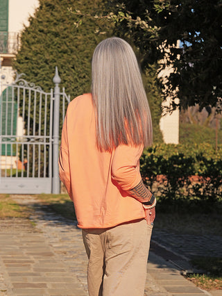 Person mit Rücken zur Kamera trägt ein OFF#DLY Longsleeve Pure apricot. Lockere Passform, lange Ärmel, sichtbar mit Rosenmotiv und Schriftzug, kombiniert mit hellen Hosen und Armbändern.