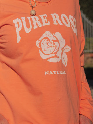 Person trägt das OFF#DLY Longsleeve Pure apricot mit einem weißen Rosenprint und den Worten PURE ROSE und NATURAL. Ein beaded Anhänger ist am Kragen sichtbar.