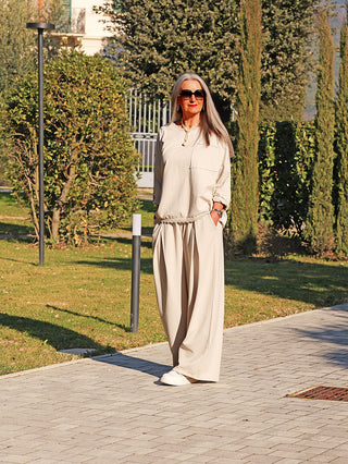 Frau trägt OFF#DLY Longsleeve Liv eisgrau, locker fallend mit Brusttasche. Steht auf einem Gartenweg, kombiniert mit weiten Hosen und Sonnenbrille, in sonniger Parkkulisse.