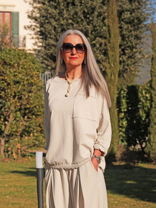 Eine Frau trägt das OFF#DLY Longsleeve Liv eisgrau, eine locker fallende, moderne Passform mit Brusttasche, kombiniert mit weiten Hosen, Perlenkette und Uhr, in einem gepflegten Garten.