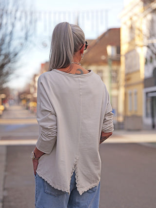 Person von hinten in OFF#DLY LS Fiona beige, einem lässigen Longsleeve mit V-förmigem Rückenschlitz und Spitzensaum, kombiniert mit locker sitzenden Jeans auf einer ruhigen Straße stehend.