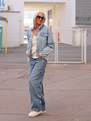 Frau mit grauen langen Haaren trägt True Religion Ruby Volume Denim: lockere, hochgeschnittene Jeans im Five-Pocket-Stil, verdeckter Reißverschluss, lässiger Look, kombiniert mit einer hellen Jeansjacke und weißen Sneakers.