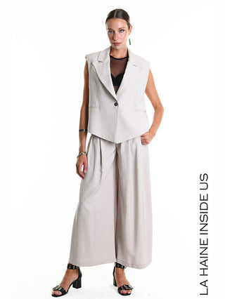 La Haine Gilet 5282 beige: Modell trägt ärmelloses, beige Gilet mit Knopfverschluss und Reverskragen über schwarzem Crop-Top, kombiniert mit weiten Hosen. Silberne Schmuckdetails und schwarze Sandalen vervollständigen den Look.