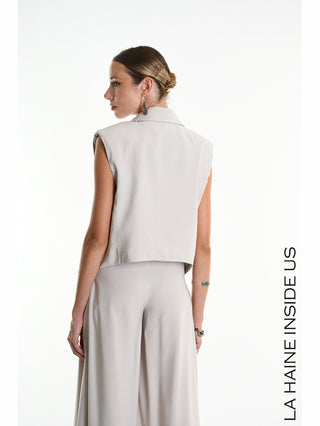 Frau trägt La Haine Gilet 5282 beige, ein ärmelloses, leicht gekürztes Gilet mit klassischem Reverskragen und weitem Bein, im Profil vor weißem Hintergrund. Elegantes und minimalistisches Design.