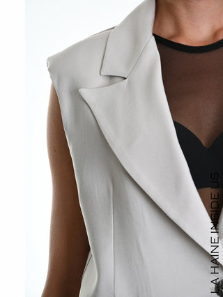 Person mit La Haine Gilet 5282 beige, ärmelloser Blazer mit klassischem Reverskragen über schwarzem Mesh-Top, enthüllt schwarze BH-Konturen. Elegantes, minimalistisches Design mit Schulterpolstern und Ein-Knopf-Verschluss.