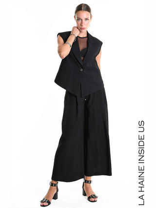 La Haine Gilet 5282 black: Modell trägt ärmelloses, schwarzes Gilet mit Ein-Knopf-Verschluss, über schwarzem Mesh-Top und weiten, schwarzen Hosen; kombiniert mit schwarzen Sandalen und silbernem Schmuck.