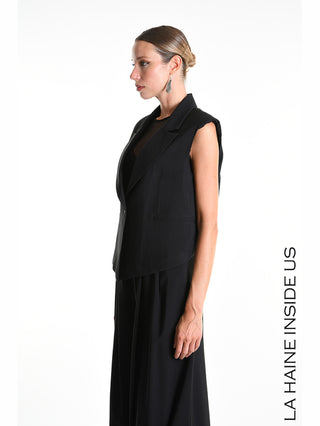 La Haine Gilet 5282 black: Modell mit schwarzem, ärmellosem Blazer-Gilet, Reverskragen und Ein-Knopf-Verschluss, kombiniert mit schwarzer, weiter Hose, präsentiert sich im Studio mit glattem Dutt und Einzelohrring.