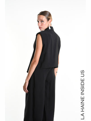 La Haine Gilet 5282 schwarz: Eine Frau trägt ein ärmelloses, schwarzes, kurzes Gilet mit weitem Schnitt, dezenten Schulterpolstern und Reverskragen, kombiniert mit schwarzen, weiten Hosen.