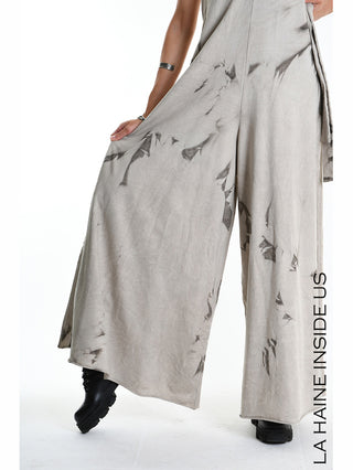 Das Bild zeigt den La Haine Jumpsuit W5296 Fango, einen weit geschnittenen Palazzo-Jumpsuit mit abstraktem Print und tiefen Seitenschlitzen, kombiniert mit schwarzen Stiefeln und einem silbernen Armreif.