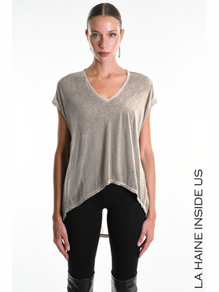 Ein Model trägt das La Haine Tee Overs. Asimetric Fango, ein asymmetrisches, locker fallendes V-Neck-Top, kombiniert mit schwarzen Leggings und kniehohen Stiefeln, vor einem schlichten Hintergrund.