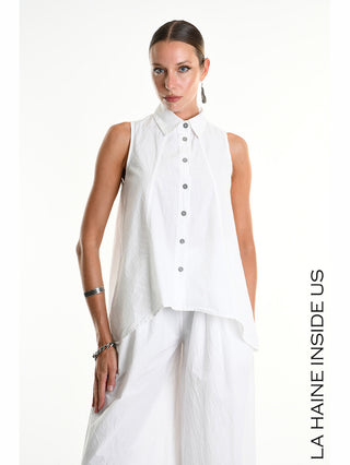 Frau trägt La Haine Shirt 5267 bianco, ärmellose asymmetrische Bluse mit geknitterter Optik und klassischem Kragen, kombiniert mit weiten Hosen. Dezenter Silberschmuck ergänzt den modernen Look.