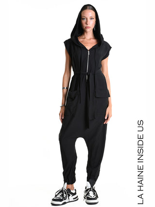 La Haine Jumpsuit M5348 black: Frau trägt einen schwarzen, ärmellosen Jumpsuit mit Kapuze, Reißverschluss, Bindegürtel, Taschen und elastischen Beinabschlüssen, kombiniert mit klobigen schwarz-weißen Sneakers.