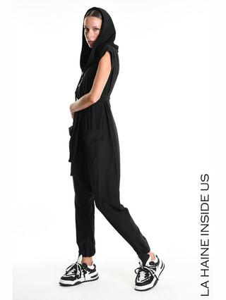 Modell in La Haine Jumpsuit M5348 black mit drapierter Kapuze, tiefem Schritt und elastischen Beinabschlüssen, kombiniert mit klobigen schwarzen Sneakers, vor weißem Hintergrund.