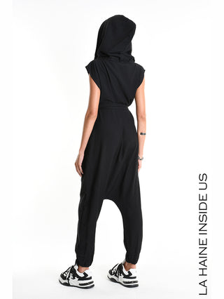 La Haine Jumpsuit M5348 black: Rückansicht einer Frau im schwarzen, ärmellosen Jumpsuit mit Kapuze und tiefem Schritt. Elastische Knöchelabschlüsse und schwarze Sneakers ergänzen den urbanen Look. Markenname rechts.