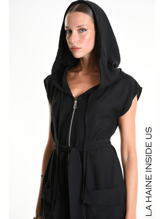 Model trägt La Haine Jumpsuit M5348 black mit Kapuze, Reißverschluss, Bindeschärpe und aufgesetzten Taschen. Der Jumpsuit hat einen tiefen Schritt und elastische Beinabschlüsse für einen urbanen Look.