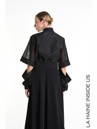 La Haine Bomber 5361 black: Rückenansicht einer schlanken Frau im verkürzten schwarzen Bomber mit abnehmbaren Tüllärmeln und elastischem Bund, kombiniert mit einem langen, fließenden Rock oder weiten Hosen.