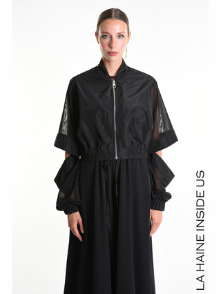 La Haine Bomber 5361 black mit abnehmbaren Tüllärmeln und Cut-Out-Details, trägt die Model auf einem weißen Hintergrund. Der Bomber hat einen Reißverschluss und eine elastische, gesmokte Taille.