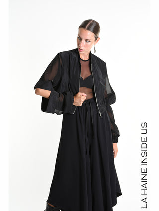 Model trägt La Haine Bomber 5361 black mit abnehmbaren Tüllärmeln, Cut-Out-Details und elastischen Bündchen. Kombiniert mit schwarzem Bralette, Rock und einem Drop-Ohrring, steht sie vor weißem Hintergrund.