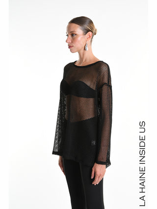 Frau in La Haine Sweater 5344 schwarz, einem grobmaschigen, transparenten Netzoberteil mit langen Ärmeln und seitlichem Schlitz, über schwarzem Bandeau und Hose, mit zurückgekämmtem Haar und Ohrringen.