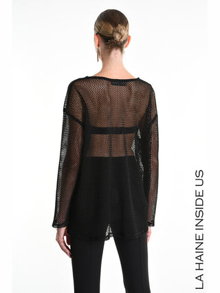 La Haine Sweater 5344 black: Rückansicht einer Frau im grobmaschigen, transparenten schwarzen Netz-Top mit langen Ärmeln und Seitenschlitz, über schwarzem Unterteil, präsentiert modernen, lässigen Layering-Look.