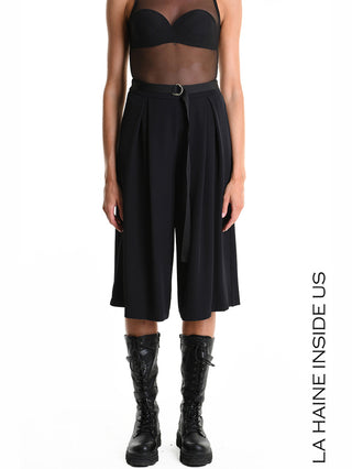 Modell trägt La Haine Bermuda LW5322 black aus fließendem Techno-Material, kombiniert mit schwarzem Mesh-Top und kniehohen Schnürstiefeln. Die Bermuda zeichnet sich durch weite Silhouette und Bügelfalten aus.