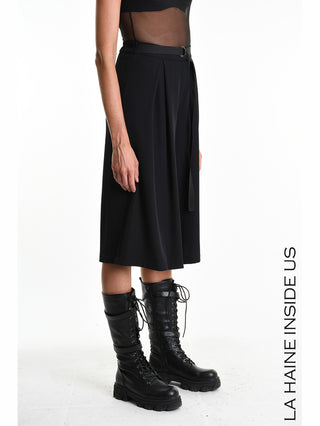 Das Bild zeigt die La Haine Bermuda LW5322 black, eine weite, knielange Bermuda aus fließendem Techno-Material, getragen mit hohen, geschnürten Stiefeln und einem transparenten schwarzen Oberteil.