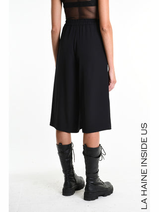 Modell von hinten in La Haine Bermuda LW5322 black: Weite, schwarze High-Waist-Culottes aus fließendem Techno-Material mit elastischem Bund, kombiniert mit schwarzen Schnürstiefeln und einem transparenten, ärmellosen Top.