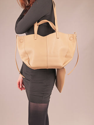 Person trägt ein schwarzes Kleid und schwarze Strumpfhosen, während sie die Regina Schrecker Tasche 5020 sand, eine beige-tan Ledertragetasche mit passender Clutch, über der Schulter trägt.