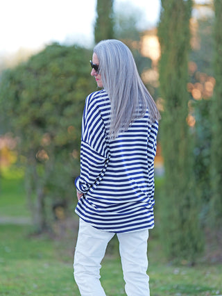 Eine Person trägt das OFF#DLY Longsleeve Lousiana marine/white mit navy-weißen Streifen und lockerem Schnitt, während sie entspannt in einer Gartenumgebung steht, größtenteils von hinten sichtbar.