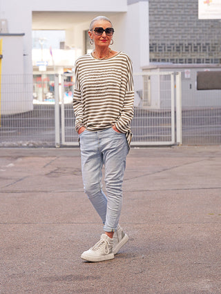 Person trägt OFF#DLY Denim Padua blue, eine schmal geschnittene, helle Jeans mit klassischem 5-Pocket-Design, kombiniert mit einem gestreiften Oberteil und Accessoires, stehend in einem urbanen Außenbereich.