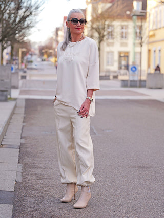 Frau trägt die OFF#DLY Fakelederhose Rimini creme, locker geschnitten, mit elastischem Bund, kombiniert mit Stiletto-Stiefeletten und passendem Oberteil, ideal für eleganten, modernen Alltagslook in urbaner Umgebung.