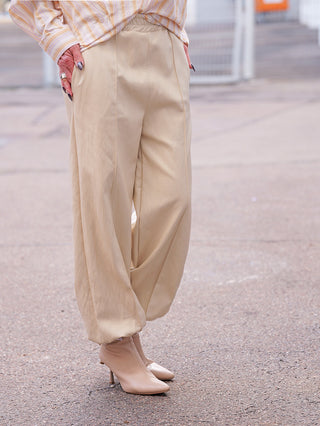 Person trägt OFF#DLY Fakelederhose Rimini creme, lockere Passform, die an den Knöcheln verjüngt ist, kombiniert mit spitzen, sockenartigen Stiefeletten; Hände in Taschen, mit silbernen Ringen und langen Nägeln.